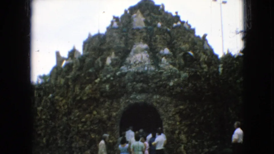 1967 Iowa home-movie frame of visitors gathered near a grotto-like religious structure with an arched opening and dense stone or foliage texture.