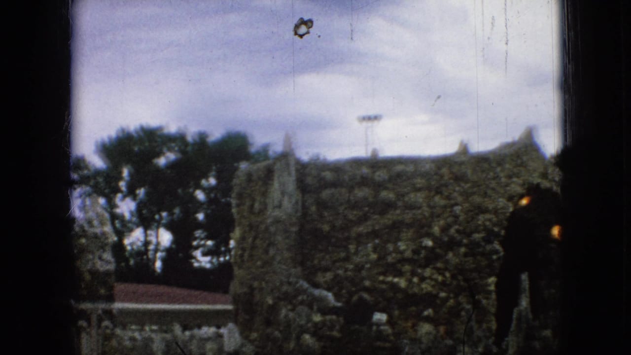 1967 Iowa 8mm frame showing visitors near a rough stone church-like wall with an arched entry and cloudy sky, partially obscured by film-frame borders.
