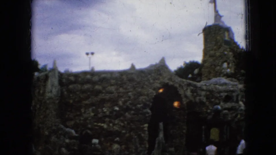 1967 Iowa home-movie frame with a stone tower topped by a cross or crucifix, arched stonework, and visitors gathered near the entrance.