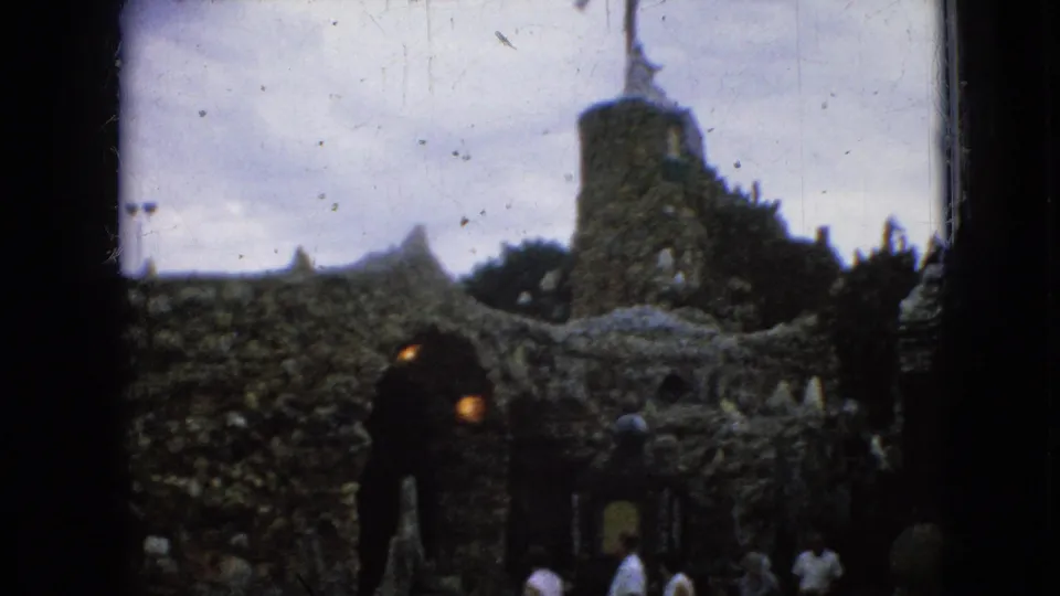 1967 Iowa 8mm frame showing several visitors around a stone church-like entrance, with the tower and cross visible above the rough masonry.