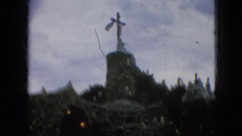 Final 1967 Iowa home-movie frame of a stone religious landmark with a cross-topped tower, rough walls, and small visitor figures below.