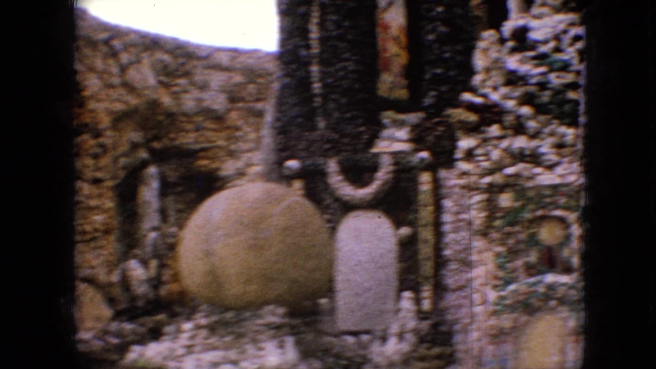 1967 Iowa 8mm frame showing a stone grotto courtyard with rounded masonry, dark vertical posts, a large circular feature, and shrine-like forms.