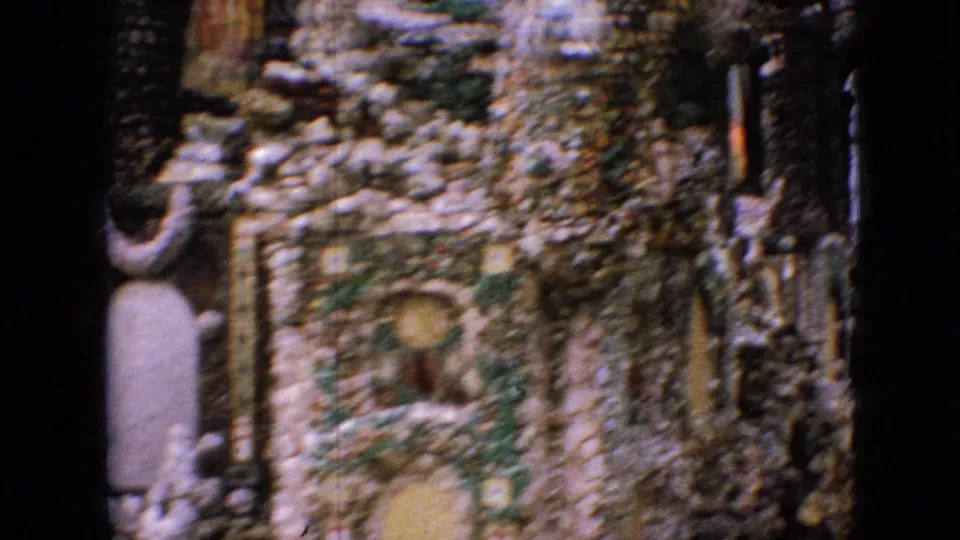 1967 Iowa 8mm frame of a grotto wall with embedded stones, arched devotional recesses, small figures, and a dark vertical post in the foreground.