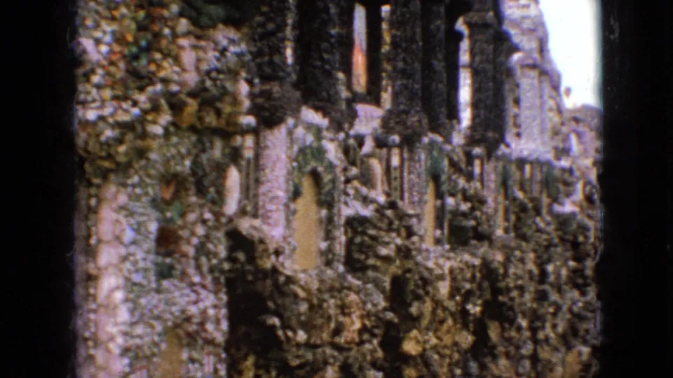 1967 Iowa 8mm frame looking along a textured stone grotto wall with small alcoves, layered masonry, and dark vertical supports beside the walkway.