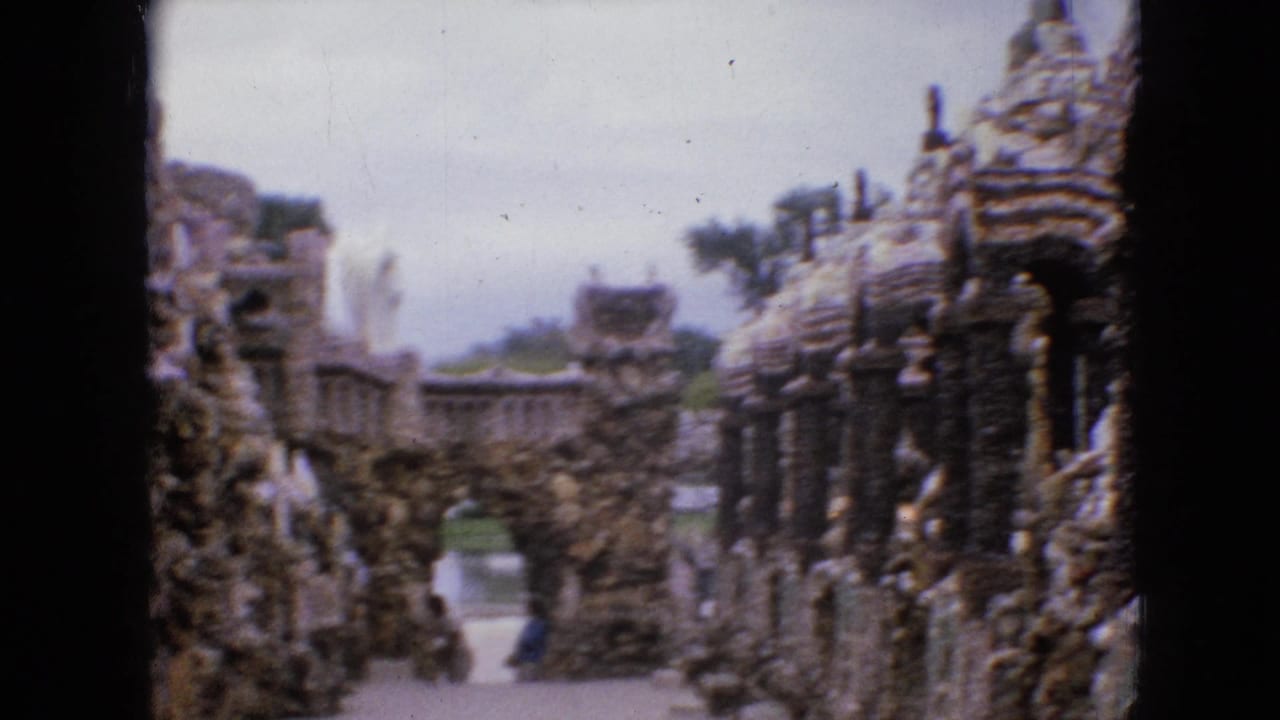 1967 Iowa 8mm home-movie frame showing a visitor's view down a path between hand-built rock and pebble structures with arches and tower forms.