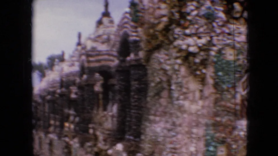 1967 Iowa home-movie frame with a close visitor-shot view of textured stone garden architecture, including pebble-covered towers, dark arch openings, and irregular masonry.