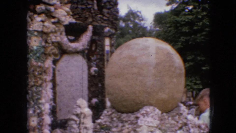 1967 Iowa home-movie frame showing a large rounded stone feature beside ornate rockwork and greenery, with a partially visible visitor at the right edge of the frame.