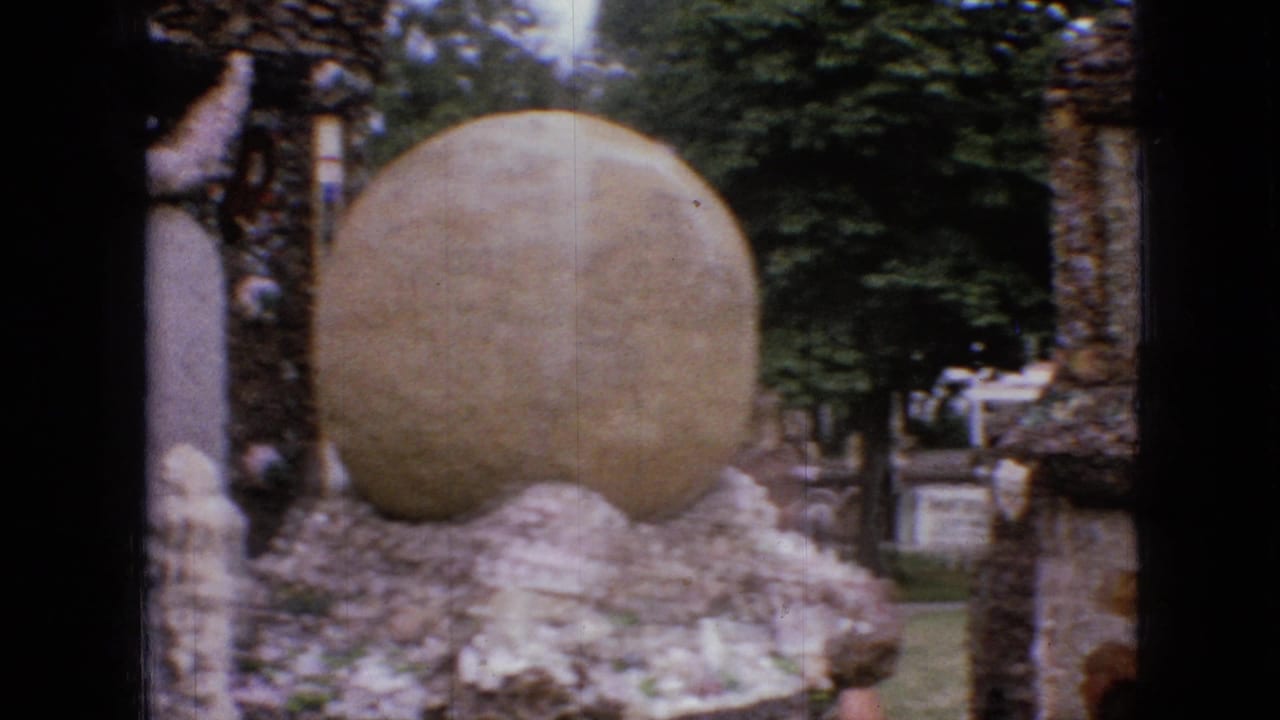 1967 Iowa 8mm frame showing a large rounded stone form set among handmade rock walls, trees, lawn, and small signs in an outdoor visitor area.