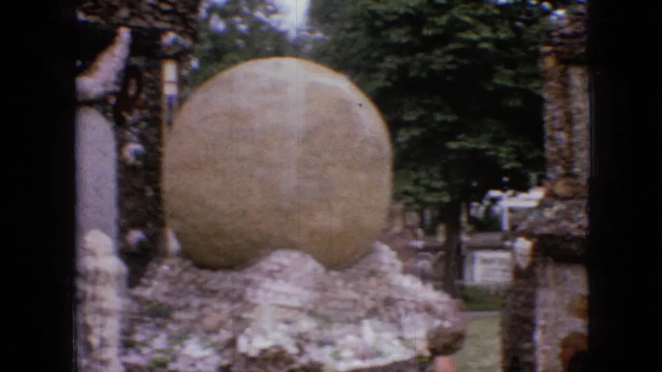 1967 Iowa 8mm frame showing a large rounded stone form set among handmade rock walls, trees, lawn, and small signs in an outdoor visitor area.