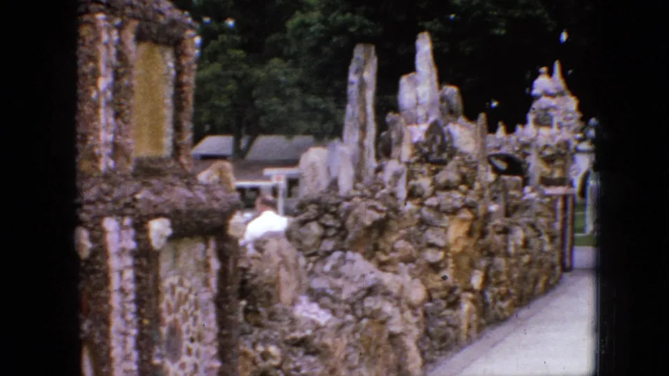 1967 Iowa 8mm frame showing a narrow walkway bordered by rough stonework, vertical sculptural forms, a pebble-textured wall, trees, and a visitor in a white shirt.