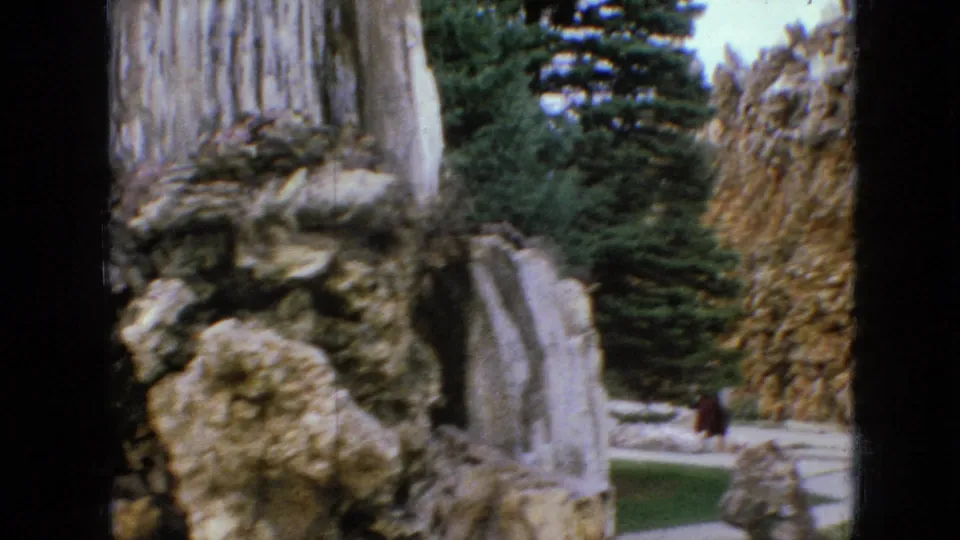 1967 Iowa home-movie frame with a close view of rough handmade stone formations, pale vertical rock surfaces, evergreen trees, lawn, and a path or open landscaped area.