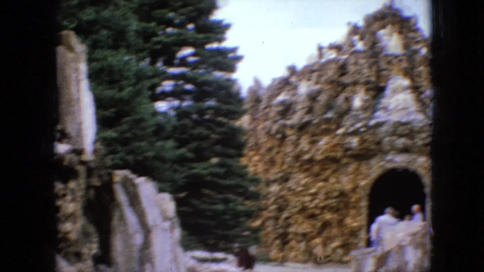 1967 Iowa 8mm frame showing a rough stone grotto with an arched opening, evergreen trees, and distant visitors near the entrance.