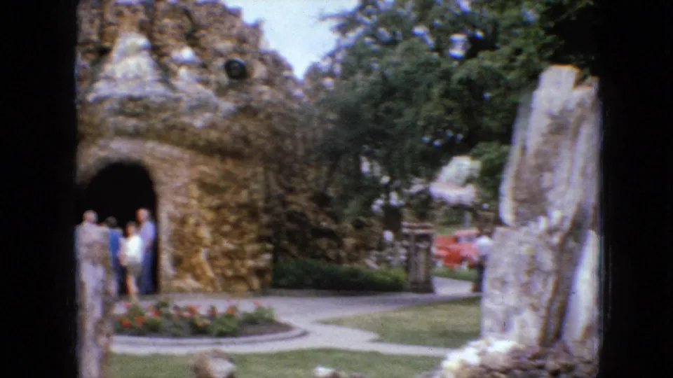 1967 Iowa 8mm frame looking across a landscaped park path toward a stone grotto entrance with visitors, flower beds, lawn, and upright rock slabs.