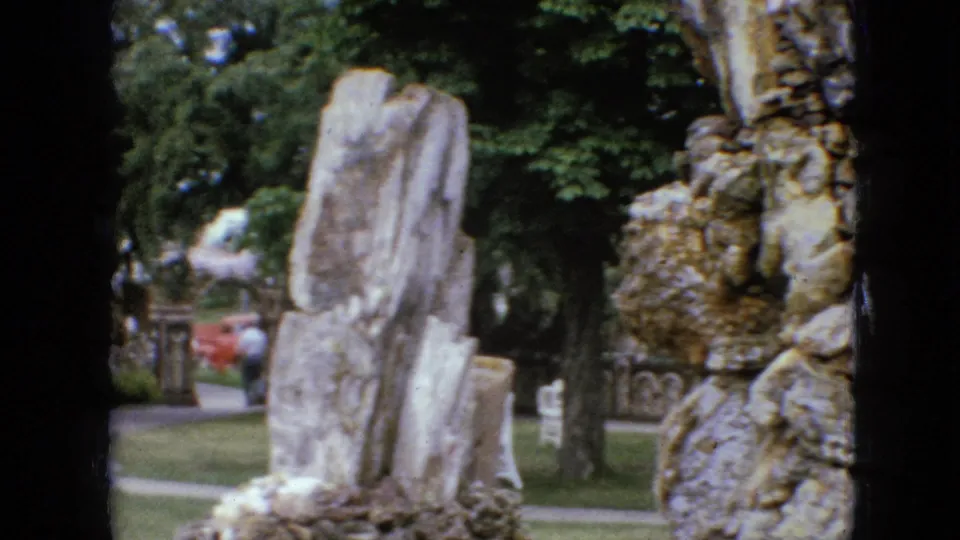 1967 Iowa home-movie frame showing upright rock slabs, trees, lawn, and a partial view of the rough stone grotto in a park setting.