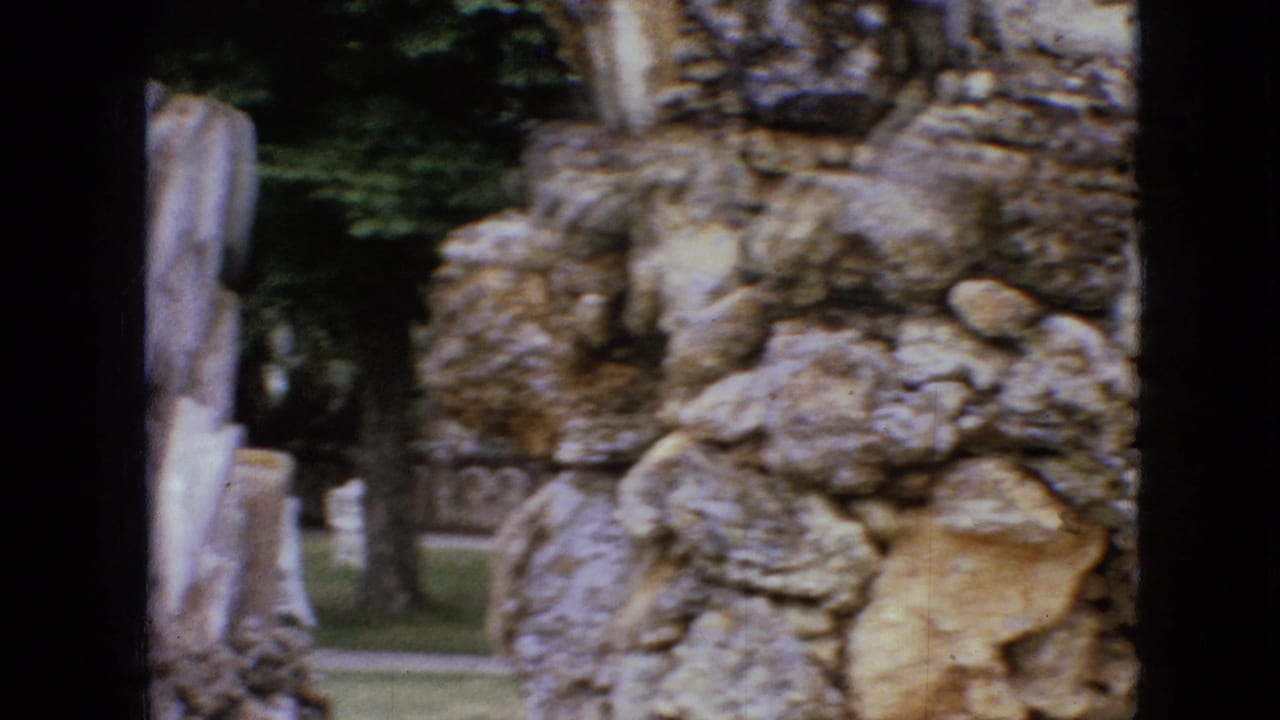 1967 Iowa 8mm frame showing a rough stone wall, pale vertical sculpted forms, and trees beyond the constructed rocky landscape.