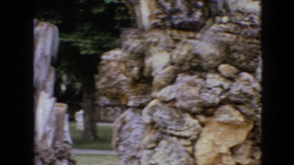 1967 Iowa 8mm frame showing a rough stone wall, pale vertical sculpted forms, and trees beyond the constructed rocky landscape.