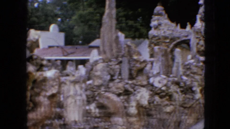 1967 Iowa 8mm frame showing a constructed rocky garden or ruin-like setting with pillar forms, pale walls, trees, and a roofed structure in the background.
