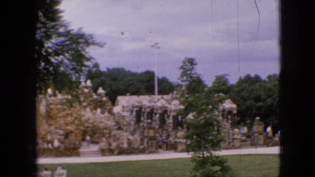 1967 Iowa 8mm frame showing a historic-looking stone structure in a park setting, seen past lawn, trees, path, and a dark window or frame divider.