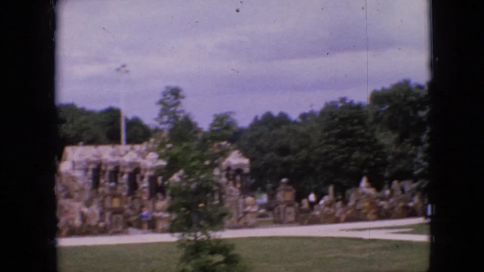 1967 Iowa home-movie frame of a historic-looking stone building or rockwork feature in landscaped grounds, with visitors walking near the path.