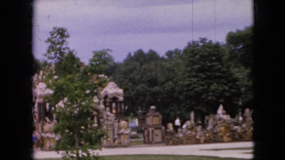 1967 Iowa 8mm still showing trees, a paved walkway, and people near elaborate stonework in a landscaped public setting.