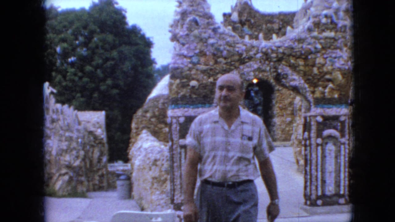 In 1967 Iowa home-movie footage, a bald middle-aged man in a striped shirt walks past rock and shell mosaic walls with an arched opening behind him.