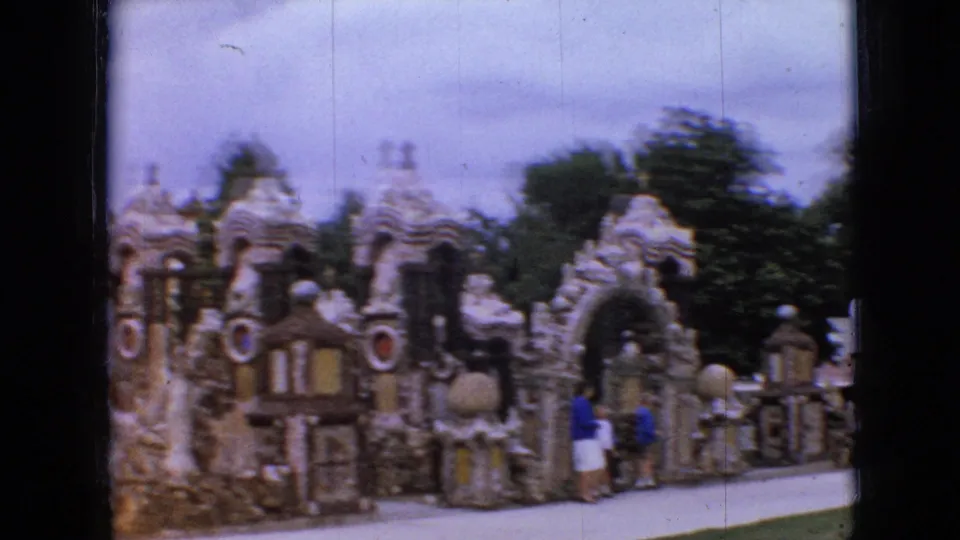 Wide 1967 Iowa tourist frame showing visitors in 1960s clothing walking along a path beside the ornate stone landmark with domes, crosses, and circular accents.