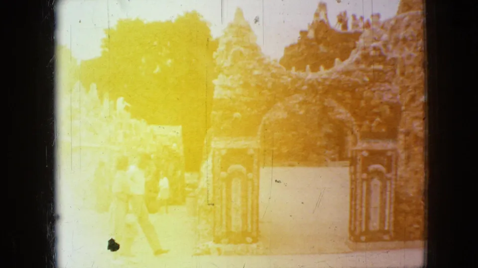 1967 Iowa 8mm frame of visitors walking by an arched stone garden entrance and decorative gate panels, with heavy yellow light leak across the scene.