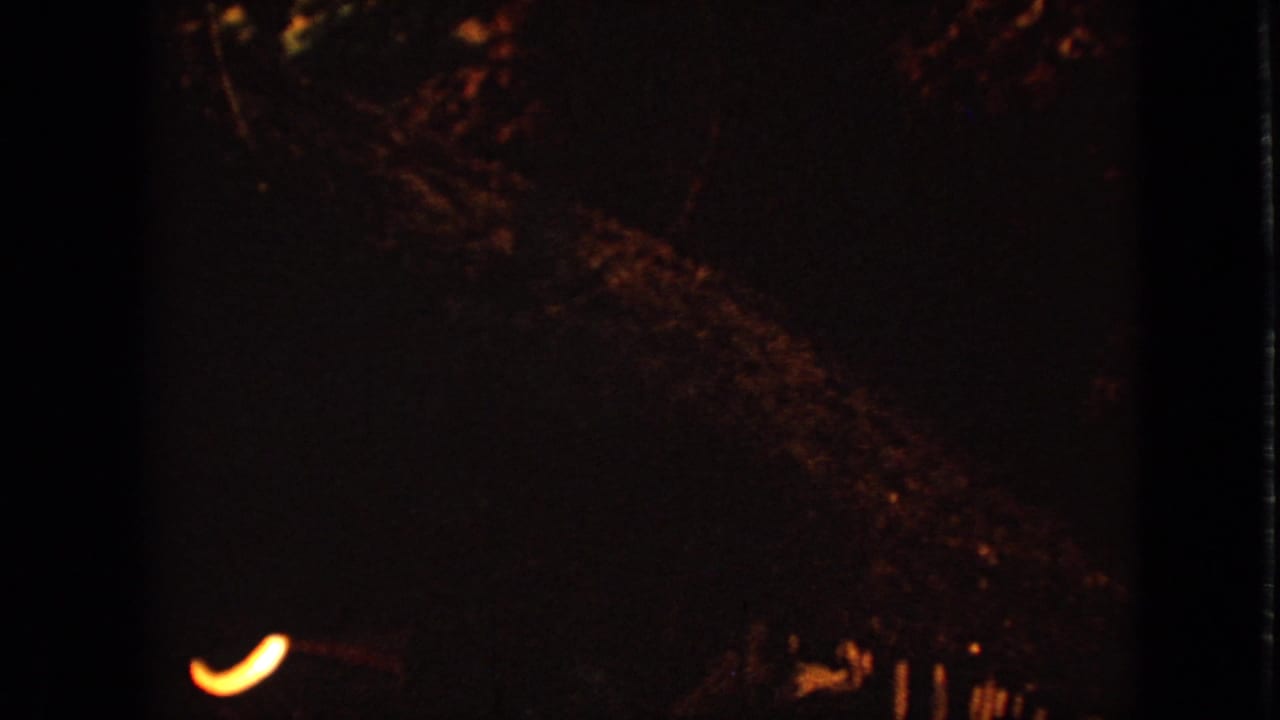 1967 Iowa 8mm frame showing a mostly dark night scene with a small curved warm light and dim surrounding foliage or holiday decoration shapes.