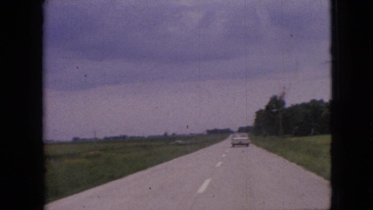1967 Iowa home-movie frame showing a two-lane rural road through open fields, with a pale car ahead under a dark, clouded sky.