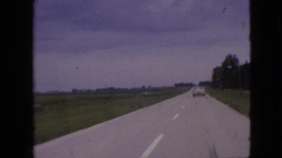1967 Iowa 8mm frame looking forward along a straight rural road, with fields, right-side trees, and a car receding into the distance.