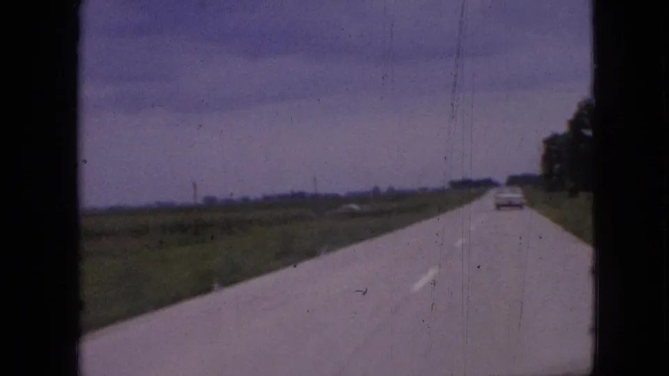 1967 Iowa home-movie view from a moving vehicle, showing a rural two-lane road bordered by fields and trees with a car ahead.