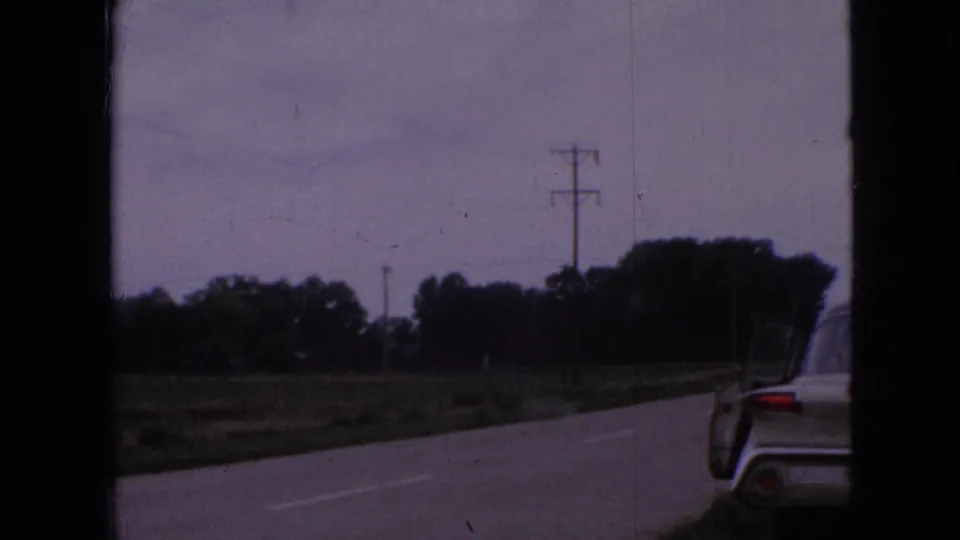 1967 Iowa 8mm frame showing a rural roadside with utility poles and trees, while part of a nearby car appears along the right edge.