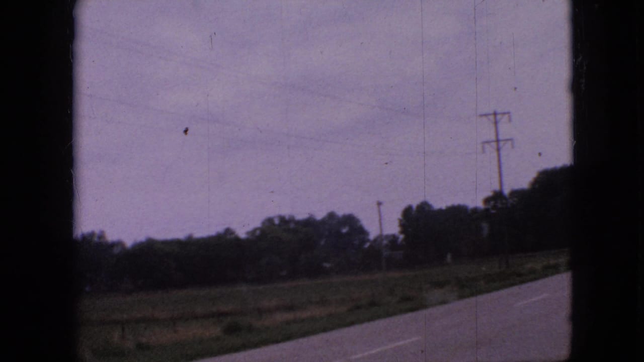 1967 Iowa 8mm frame showing a rural paved road, grassy farmland, trees, and utility poles seen from a moving vehicle.