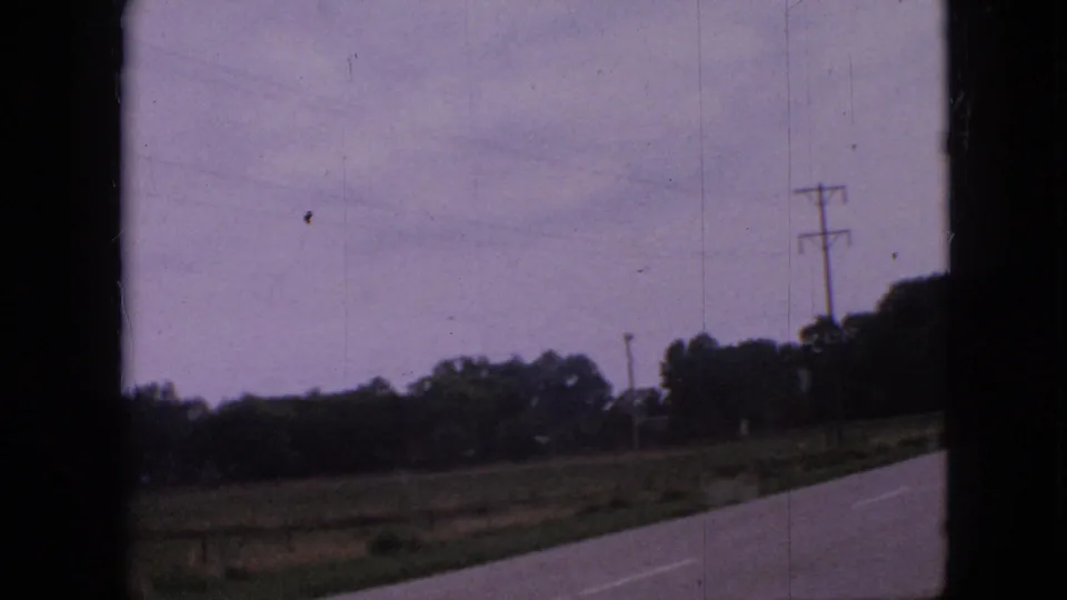1967 Iowa 8mm frame showing a rural paved road, grassy farmland, trees, and utility poles seen from a moving vehicle.