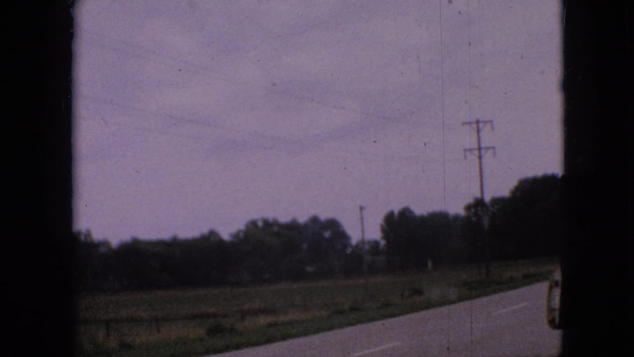 A passing-vehicle view of a 1967 Iowa rural road with open fields, utility poles, and a tree line in soft 8mm color.