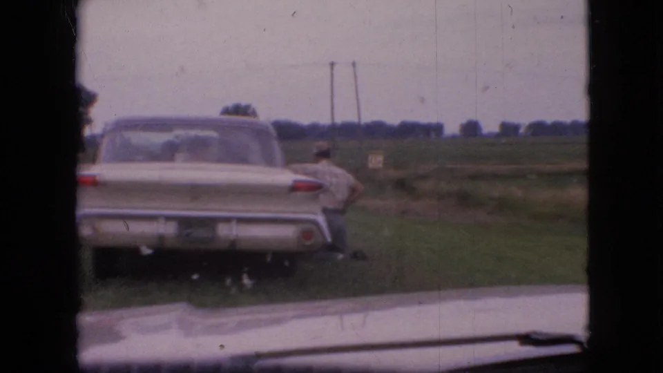 A cream-colored classic car is parked on a grassy Iowa roadside in 1967, with an adult man partly visible near the passenger side and open fields beyond.