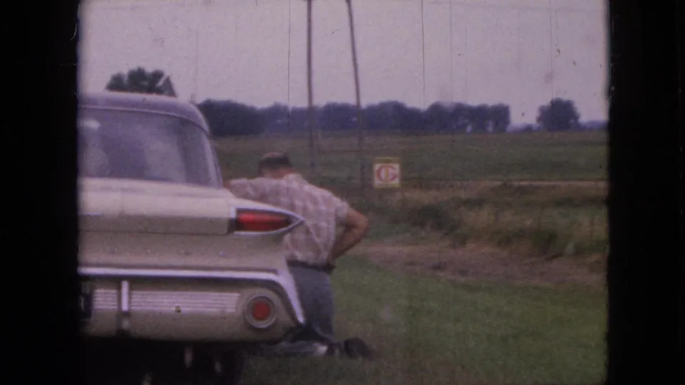 An adult man in a short-sleeve shirt appears crouched or kneeling beside a cream-colored classic car on a rural Iowa roadside in 1967.