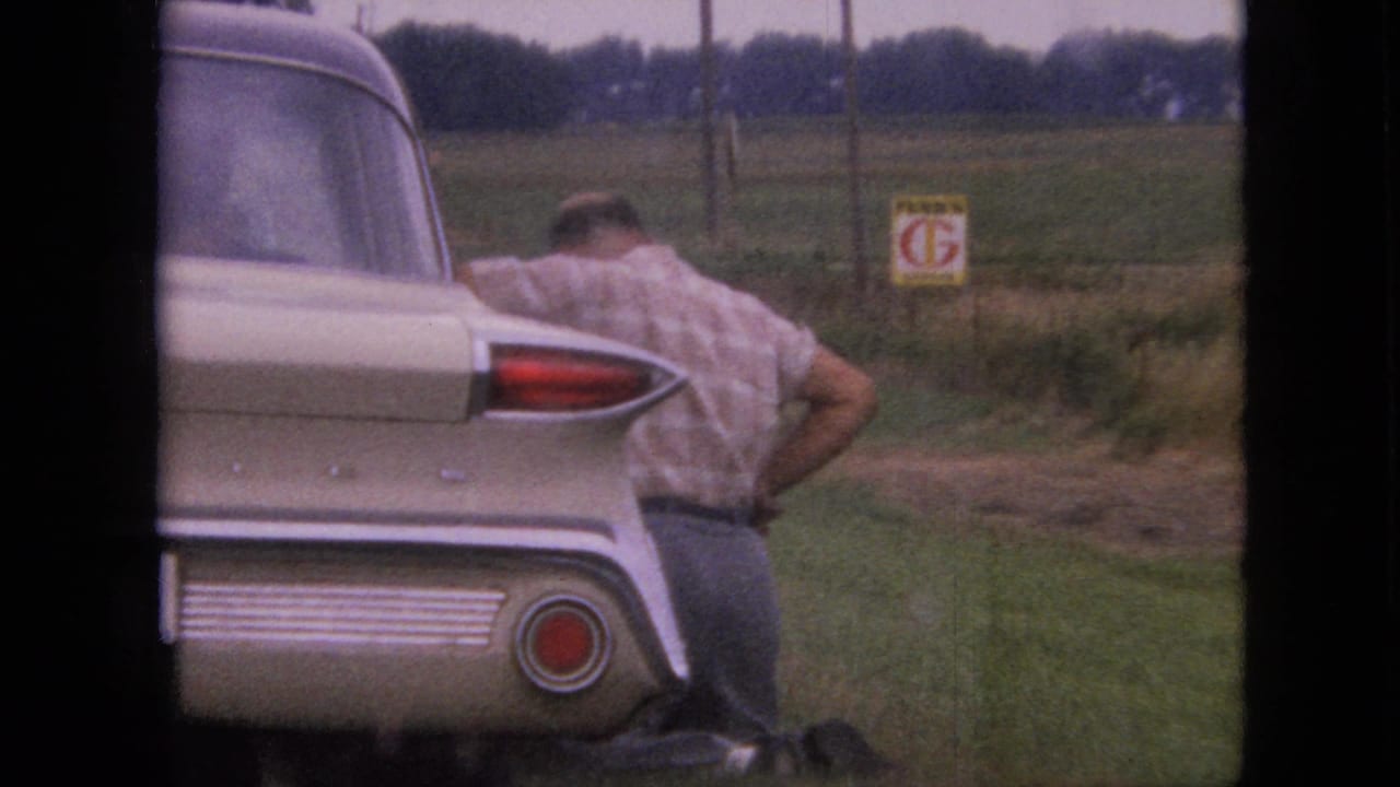 Man leaning over the rear of a cream-colored classic car beside a rural field.