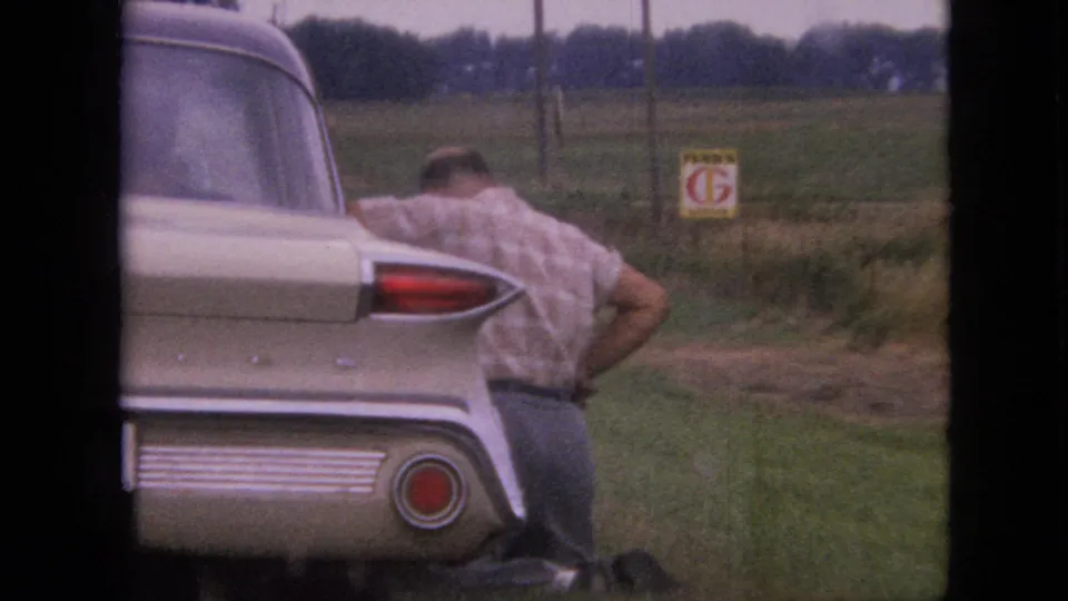 In Iowa home-movie footage from 1967, a man in a plaid shirt leans near the rear of a cream-colored classic car parked on a grassy roadside by fields and fence posts.