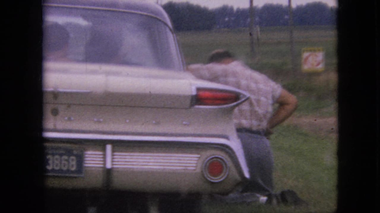 Rear of a cream-colored classic car with a man bent over beside it near a field.