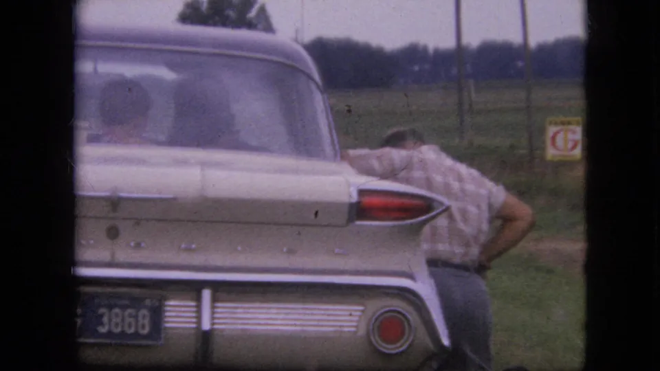 The rear of the cream-colored car fills the frame while the man remains bent beside it, with the partial license plate, red taillight, field, and sign visible in the 1967 Iowa scene.