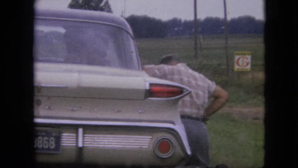 A man stands bent beside the rear of a cream-colored classic car on a grassy rural roadside in Iowa, with fields, fence posts, and a small sign behind the vehicle.