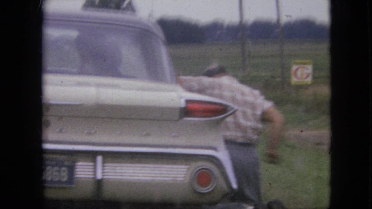 In 1967 Iowa home-movie footage, a man is partly obscured near the rear of a light-colored tailfin-era sedan parked beside a grassy rural road.