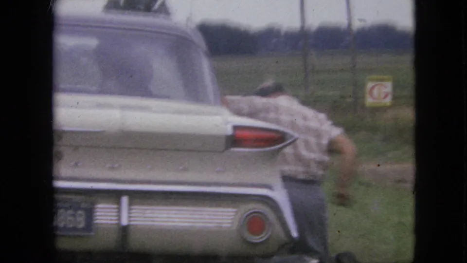 In 1967 Iowa home-movie footage, a man is partly obscured near the rear of a light-colored tailfin-era sedan parked beside a grassy rural road.