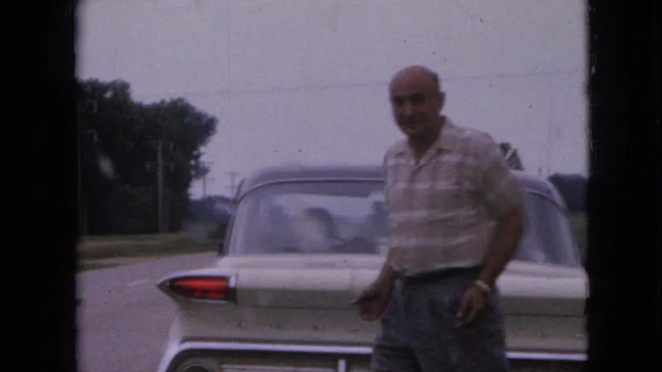 The man faces the camera near the back of the light-colored sedan, with the rural Iowa road and trees visible behind him.