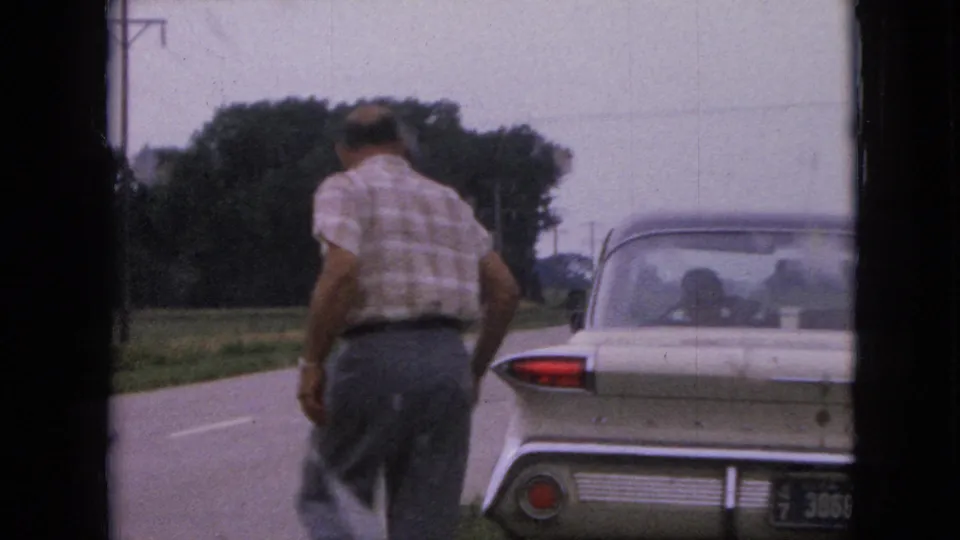 A rear view shows the man walking beside the parked tailfin sedan along the grassy edge of a rural Iowa road.