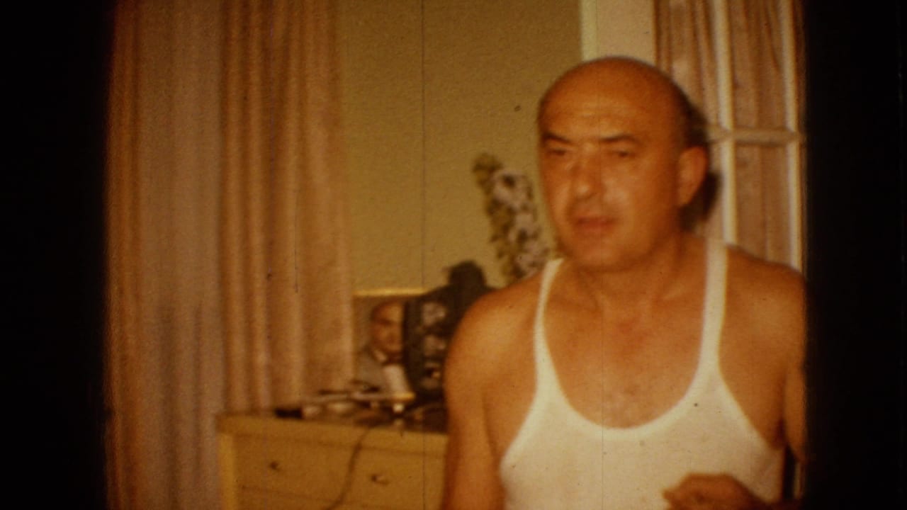 In 1967 Iowa home-movie footage, a bald adult man in a white tank top faces the camera inside a warmly lit room with beige curtains, a dresser, and framed photographs behind him.