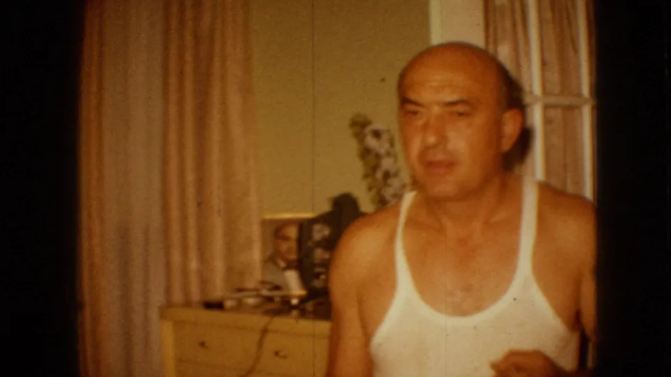 In 1967 Iowa home-movie footage, a bald adult man in a white tank top faces the camera inside a warmly lit room with beige curtains, a dresser, and framed photographs behind him.