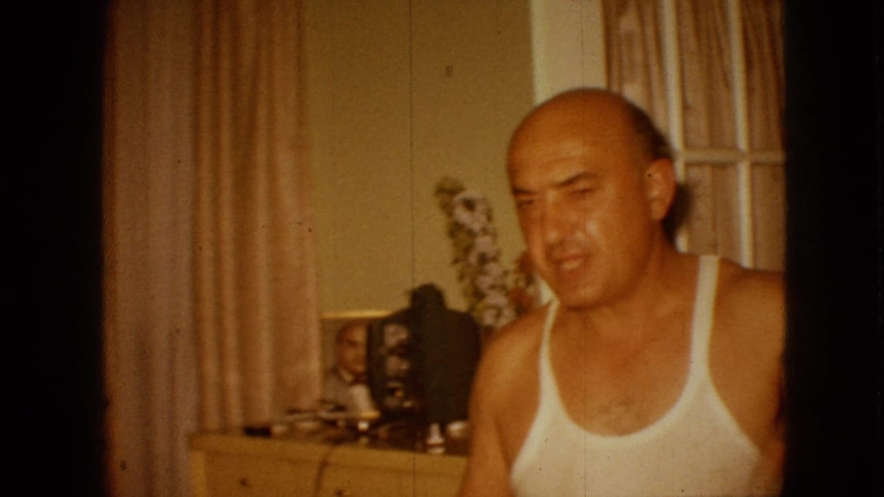 Man in a white tank top looks downward beside beige curtains and a dresser with framed photographs.