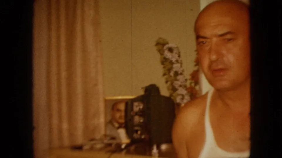 A closer 1967 Iowa frame shows the bald man near the right edge, his face and shoulder filling much of the shot while curtains, framed photos, and a floral arrangement remain in the background.